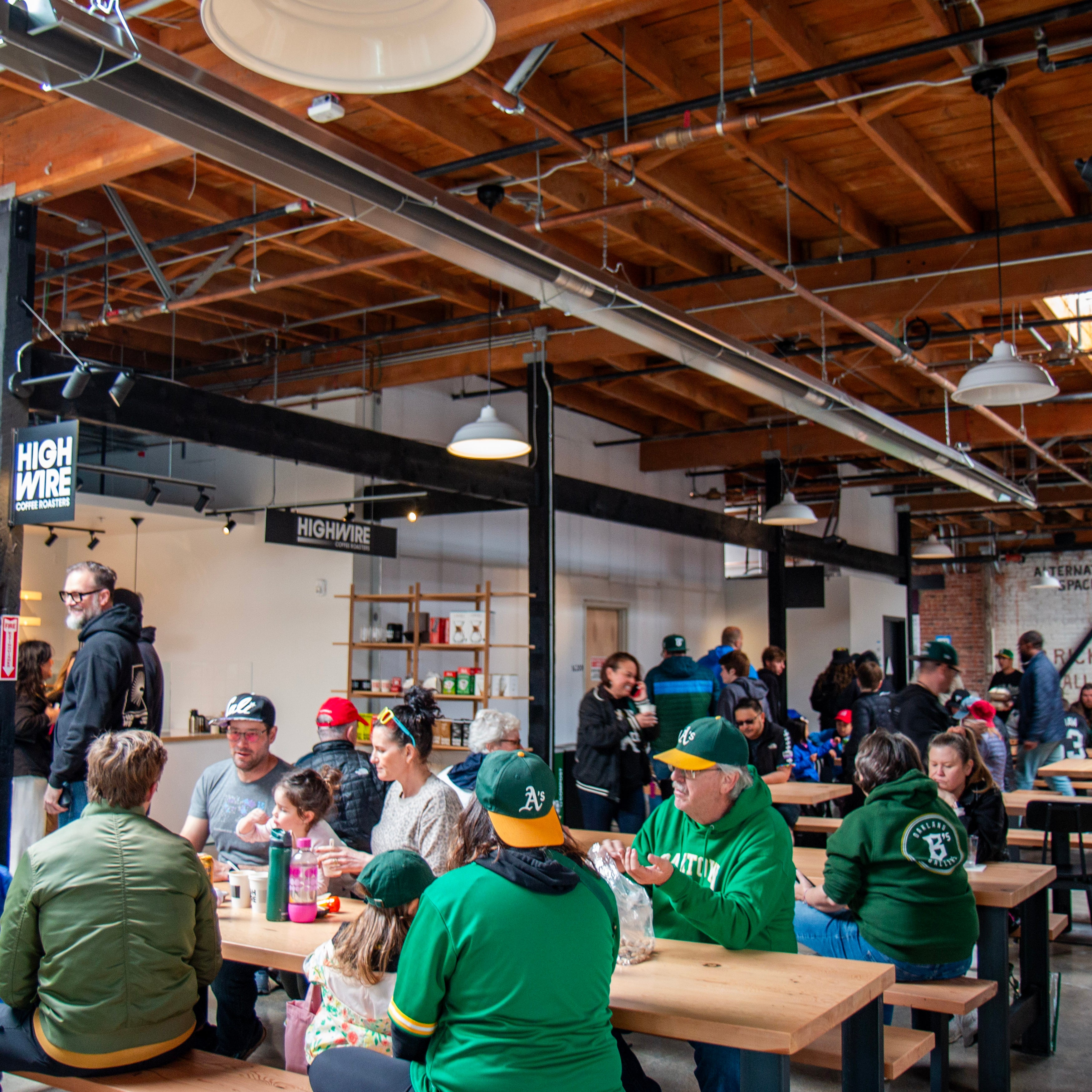 A busy interior scene at Highwire's coffee shop in Prescott Market Hall. Many of the customers are sporting Oakland A's merchandise. The owners chat beneath a Highwire sign.
