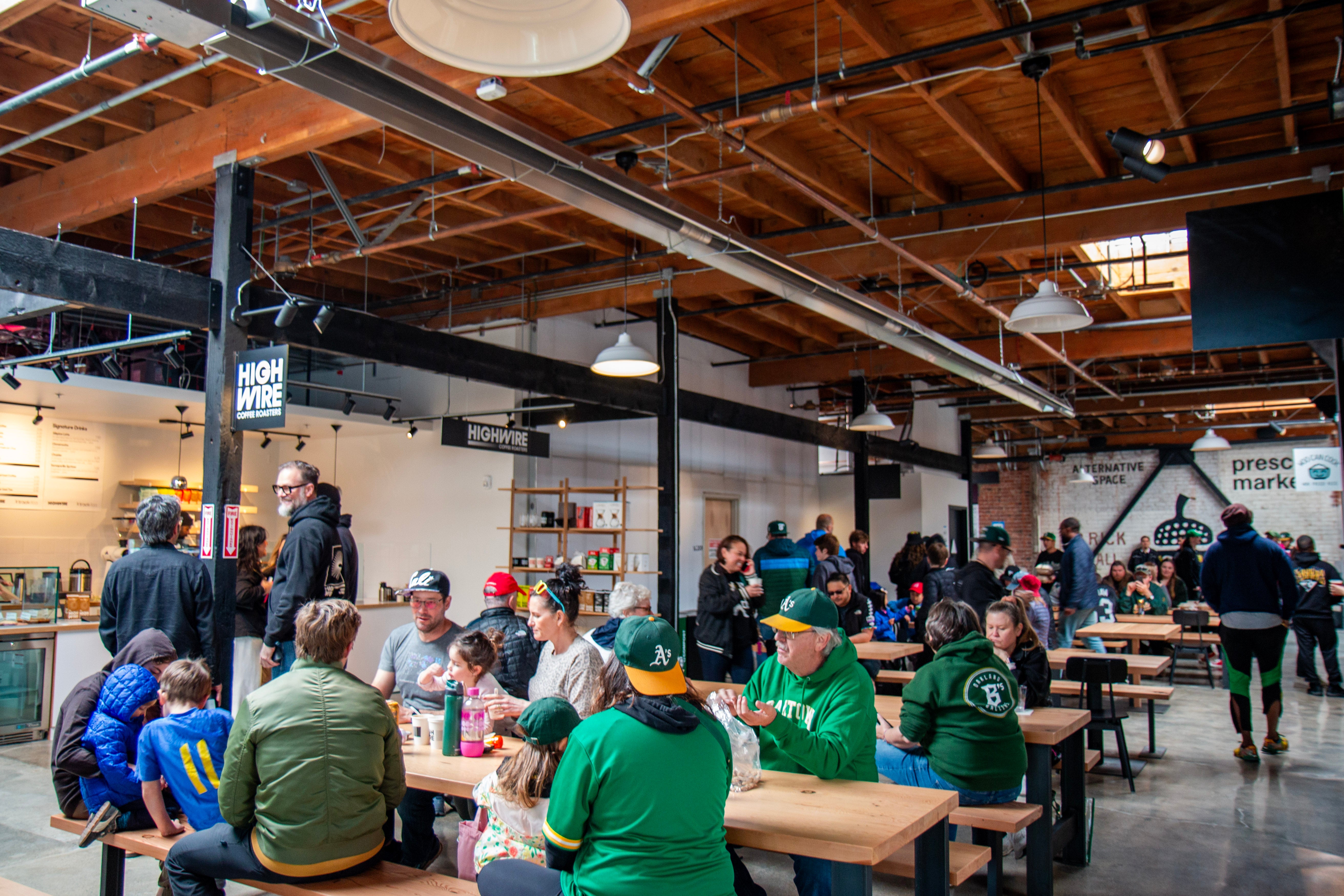 A busy interior scene at Highwire's coffee shop in Prescott Market Hall. Many of the customers are sporting Oakland A's merchandise. The owners chat beneath a Highwire sign.